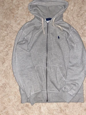 Grå zip hoodie från Polo Ralph Lauren - Snygg grå hoodie med dragkedja från Polo Ralph Lauren. Klassisk huva med vita snören och mörkblå broderad logga på bröstet. Mjuk bomullsmix och två fickor framtill. Perfekt för en chill och avslappnad stil.
