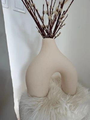 Beige modern vas i keramik - Säljer en beige vas i keramik med unik, bågformad design. Perfekt som inredningsdetalj för att ge rummet en modern och stilren känsla. Passar snyggt med torkade blommor eller kvistar.