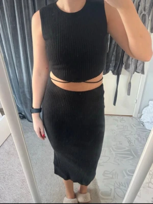 Svart ribbat set med omlottband - Snyggt svart ribbat set med crop top och omlottband i midjan. Kjolen är midilång med slits bak och snörning i ryggen för extra detalj. 