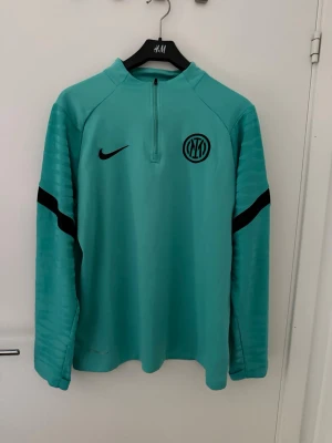 Långärmad fotbollströja - L men passar även M. Snygg turkos långärmad träningströja från Nike med Inter-logga på bröstet. Tröjan har halv dragkedja framtill och svarta detaljer på ärmarna. Perfekt för träning eller uppvärmning. Tillverkad i ett lätt och funktionellt material.