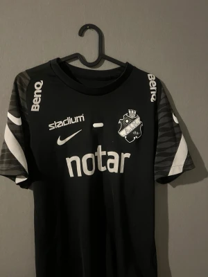 Svart AIK matchtröja Nike - Säljer en svart AIK matchtröja från Nike med vita loggor och tryck från sponsorer som Notar, Stadium och BenQ. Tröjan har korta ärmar med gråmönstrade detaljer och klubbmärke på bröstet. Perfekt för dig som älskar fotboll och AIK.