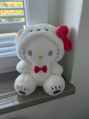 Söt vit Hello Kitty mjukisdjur - Supergulligt mjukisdjur som föreställer Hello Kitty i en vit dräkt med röd rosett på huvudet och en liten röd fluga. Plyschmaterialet är mjukt och fluffigt, och tassarna har bruna detaljer. Perfekt som present eller dekoration. 💗☺️