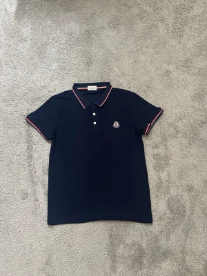 Marinblå pikétröja från Moncler - Snygg marinblå pikétröja från Moncler med klassisk krage och knappar framtill. Tröjan har röda och vita detaljer på krage och ärmslut samt Moncler-logga broderad på bröstet. Perfekt för en stilren och sportig look.