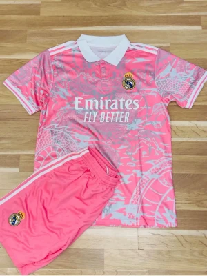 Rosa Real Madrid fotbollskit - Säljer ett rosa Real Madrid fotbollskit med matchande tröja och shorts. Tröjan har vit krage, vita detaljer och klubbmärke på bröstet samt Emirates Fly Better-tryck. Setet har ett unikt mönster i ljusrosa och vitt, perfekt för träning eller match.
