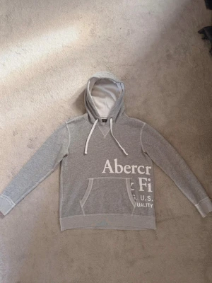 Grå hoodie från Abercrombie & Fitch - Snygg grå hoodie med vit text från Abercrombie & Fitch. Hoodien har en stor magficka, vita dragsnören och tryck både fram och bak. Tillverkad i mjukt material och har en klassisk passform med ribbade muddar vid ärmar och nederkant.