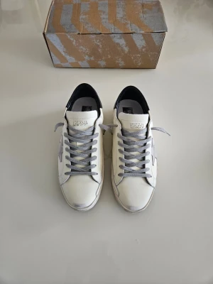 Golden goose skor  - Snygga vita sneakers med grå stjärndetalj på sidan och grå skosnören. Skorna har svart hälparti och perforerade detaljer på plösen. Tillverkade i läder med en klassisk låg siluett som passar till många olika stilar.                                                                               Pris kan diskuteras.                                                            