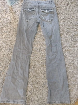 Grå bootcut jeans med kontrastsömmar - Säljer ett par grå jeans med bootcut-modell och tydliga vita kontrastsömmar. Jeansen har två bakfickor med lock och knappdetaljer samt bälteshällor. Materialet är klassisk denim i bomull. Perfekta för en avslappnad och trendig look.