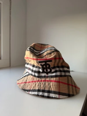 Beige bucket hat med rutigt mönster - Snygg bucket hat i klassiskt beige, svart, vitt och rött rutigt mönster. Hatten har ett broderat B monogram framtill och är tillverkad i ett tunt, lätt tyg som känns luftigt. Perfekt accessoar för att lyfta din stil med en ikonisk look.