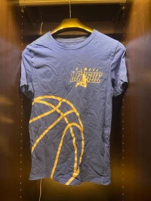 Blå t-shirt med baskettryck - Snygg blå t-shirt med gult basketboll-tryck och texten 'Global League' framtill. Klassisk rund hals och korta ärmar. Perfekt för dig som gillar sportig stil och vill sticka ut med ett coolt motiv.