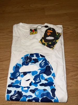 Vit BAPE t-shirt med blått camomönster - Säljer en vit t-shirt från BAPE med det klassiska apansiktet i blått camomönster på bröstet och texten 'BY BATHING APE' under. T-shirten har korta ärmar och rund hals. Perfekt för dig som gillar streetwear och vill sticka ut.