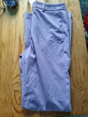 Ljuslila kostymbyxor med raka ben - Snygga ljuslila kostymbyxor med raka ben och klassisk midja. Byxorna har bälteshällor och pressveck framtill för en stilren look. Perfekta för dig som vill sticka ut med färg i din outfit.