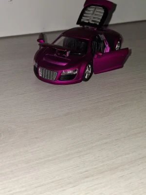 Lila modellbil Audi R8 - Säljer en detaljerad modellbil av Audi R8 i metalliclila färg. Bilen har öppningsbara dörrar, motorhuv och baklucka. Tillverkad i metall och plast med realistiska detaljer som synliga motor- och interiördelar. Perfekt för samlare eller som dekoration. Vissa plastdetaljer saknas se bilder.