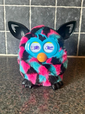 Färgglad elektronisk Furby-leksak - Säljer en elektronisk Furby med mjuk päls i svart, rosa och turkos. Den har stora svarta öron, blå plast runt ögonen och orange näbb. Ögonen är digitala och kan visa olika uttryck. Perfekt för dig som gillar retroleksaker eller samlar på Furbys.Den är varsamt använd.