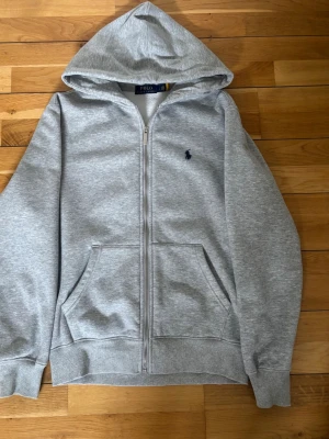 Grå zip hoodie med huva - Snygg gråmelerad hoodie från Polo Ralph Lauren med dragkedja framtill och klassisk huva. Har två stora fickor fram och en liten mörkblå logga broderad på bröstet. Tillverkad i mjukt sweatshirtmaterial, perfekt för en chill och avslappnad stil.