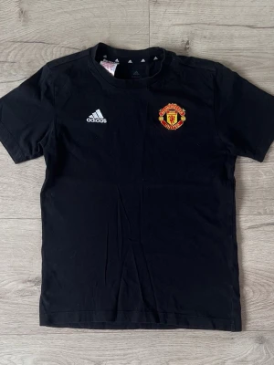 Svart Manchester United t-shirt Adidas - Svart t-shirt från Adidas med Manchester United-logga på bröstet och Adidas-logga i vitt. Klassisk rund hals och korta ärmar. Perfekt för dig som älskar fotboll och vill visa vilket lag du hejar på.