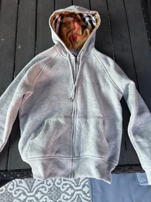 Grå zip hoodie från Burberry - Snygg grå zip hoodie från Burberry med klassiskt rutigt foder i huvan. Hoodien har dragsko i huvan, två stora fickor fram och ribbade muddar. Perfekt för dig som gillar stilrena plagg med exklusiv känsla och ikoniska detaljer. Plus aldrig använd👍🏼