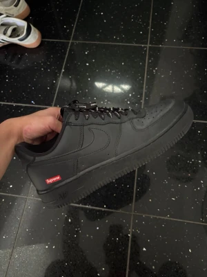 Svarta Supreme x Nike Air Force 1 - Säljer ett par svarta Supreme x Nike Air Force 1 sneakers i läder. Skorna har klassisk låg siluett, perforerad tåbox och Supreme-logga på sidan. Svarta skosnören med Supreme-detaljer och Nike Swoosh på sidorna. Perfekta för dig som gillar streetwear.