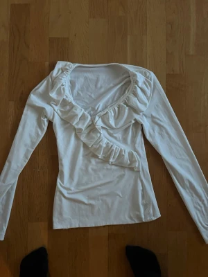 Vit långärmad topp med volang - Snygg vit långärmad topp med v-ringning och volangdetalj som går diagonalt över bröstet. Tillverkad i mjukt material, perfekt för dig som gillar en feminin och stilren look. Passar till både jeans och kjol.