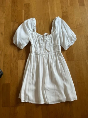 Vit kort klänning med puffärmar - Söt vit kortklänning med puffiga kortärmar och snörning framtill. Klänningen har en omlottliknande överdel och markerad midja som ger en feminin siluett. Tillverkad i ett lätt och luftigt material, perfekt för varma dagar. JÄTTE skönt material 