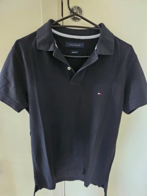 Svart pikétröja från Tommy Hilfiger - Klassisk svart pikétröja från Tommy Hilfiger med korta ärmar och liten broderad logga på bröstet. Tröjan har krage och två knappar vid halsen. Regular fit-modell i mjuk bomull som passar perfekt till jeans eller shorts.