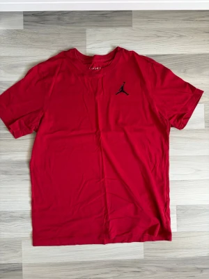 Röd Jordan t-shirt med logga - Säljer en röd t-shirt från Jordan med den klassiska svarta loggan på bröstet. T-shirten har rund hals och korta ärmar, perfekt för en sportig och avslappnad stil. Tillverkad i mjuk bomull för skön komfort.