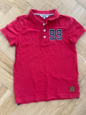 Röd pikétröja med siffran 99 - Klassisk röd pikétröja med krage och knappar framtill. Siffran 99 är tryckt på bröstet i vitt och svart. Tröjan har korta ärmar och är tillverkad i mjuk bomull, perfekt för en avslappnad stil.