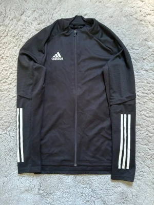 Svart träningsjacka från Adidas - Säljer en svart träningsjacka från Adidas med hel dragkedja framtill och vita ränder på ärmarna. Jackan har hög krage och är tillverkad i ett lätt, tekniskt material (AEROREADY) som andas. Perfekt för träning eller uppvärmning.