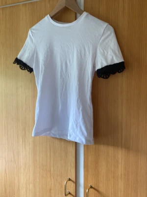Vit t-shirt med svart spetskant - Säljer en vit t-shirt med korta ärmar och snygg svart spetskant vid ärmsluten. Enkel och stilren modell som passar perfekt till jeans eller kjol. Tillverkad i mjukt material för skön känsla hela dagen.