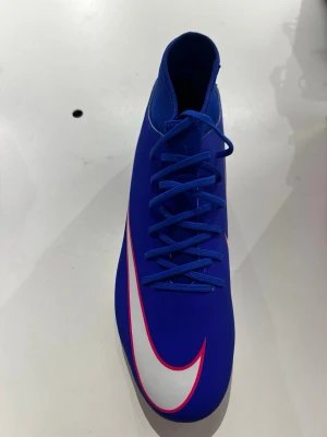 Blåa Nike fotbollsskor med hög krage - Snygga blåa fotbollsskor från Nike med hög krage och klassisk vit swoosh med rosa kant. Skorna har snörning och är tillverkade i ett syntetmaterial som ger bra passform och kontroll på planen. Perfekta för dig som vill sticka ut på fotbollsplanen.