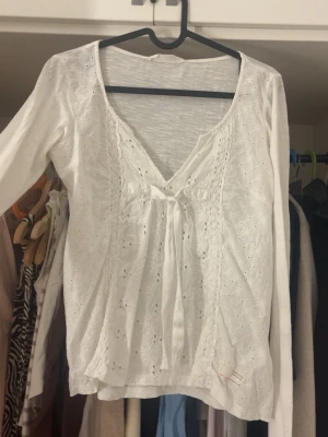 Vit blus med spets och knyt - Söt vit blus med v-ringning, spetsdetaljer och knyt framtill. Blusen har lång ärm och är dekorerad med små broderade hålmönster som ger en romantisk känsla. Perfekt att styla med jeans eller kjol för en trendig look.