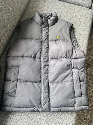Grå dunväst från lyle and scott - Snygg grå dunväst med hög krage och två stora fickor framtill. Lyle and scott väst med quiltad design som ger en puffig look. Perfekt för lager-på-lager och håller dig varm under kyliga dagar. Finns likadan väst på rea för 799kr.