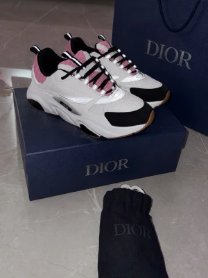 Dior vita och rosa chunky B22 - Snygga chunky sneakers från Dior i vitt, svart och rosa med mesh och läderdetaljer. Skorna har svarta och vita snören, grov sula och coola kontrastpaneler. Perfekta för dig som vill sticka ut med exklusiv streetstyle.