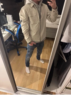 Beige overshirt C.p company - Snygg beige overshirt med dragkedja framtill och bröstficka. Har en svart patch med glasdetalj på vänster ärm som ger en cool touch. Overshirten har klassisk krage och raka ärmar. Perfekt att slänga över en hoodie för en avslappnad look.