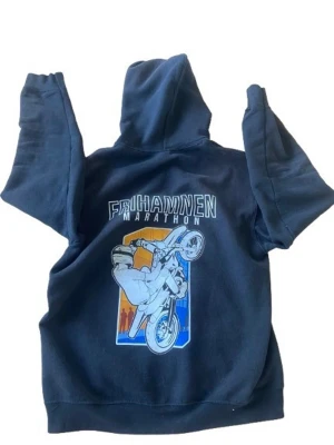 Frihamnen Marathon hoodie - Mörkblå hoodie med tryck på ryggen som visar en motorcykel och texten 'Frihamnen Marathon'. Klassisk känguruficka framtill och justerbar huva med dragsko. Skön och avslappnad passform, perfekt för dig som gillar streetwear och coola prints.