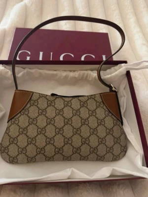 Gucci beige monogram handväska - Säljer en snygg beige handväska från Gucci med klassiskt GG-monogram i canvas och bruna läderdetaljer. Väskan har en smal axelrem och dragkedja upptill. Perfekt för dig som gillar stilrena och ikoniska accessoarer.