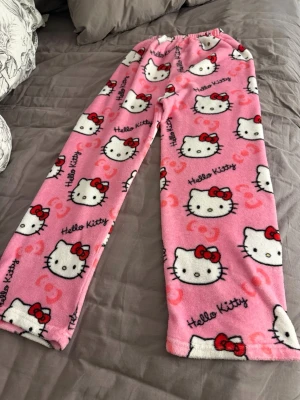 Rosa Hello Kitty pyjamasbyxor - Mysiga rosa pyjamasbyxor med Hello Kitty-motiv och röda rosetter. Byxorna har resår i midjan och är gjorda i mjuk fleece som känns superbekväm. Perfekta för chillkvällar hemma eller när du vill ha det extra cozy.