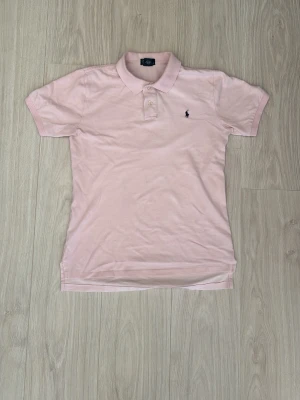 PoloRalphLauren Piké - Säljer en ljusrosa pikétröja med klassisk krage och två knappar framtill. Tröjan har korta ärmar och en liten broderad logga på bröstet. Perfekt för en avslappnad men ändå stilren look.