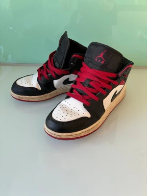 Air Jordan 1 Mid svart/vit/röd - Säljer ett par Air Jordan 1 Mid sneakers i svart, vitt och rött läder med röda skosnören och klassisk Nike Swoosh på sidan. Skorna har perforerad tåbox och ikoniska Air Jordan-loggan på sidan. Perfekt för dig som gillar streetwear och snygga detaljer.