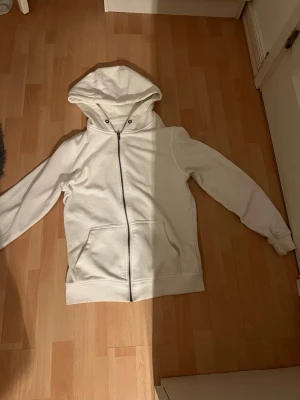 Vit hoodie från Urban Classics - Säljer en stilren vit hoodie från Urban Classics i storlek S. Hoodien har dragkedja framtill, huva med snörning och är tillverkad i ett mjukt bomullsmaterial. Perfekt för en avslappnad och clean look.