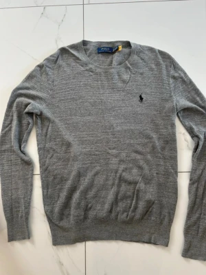 Grå stickad tröja Polo Ralph Lauren - Snygg grå stickad tröja från Ralph Lauren med marinblå logga på bröstet. Tunn och perfekt till lager på lager och enkel att matcha. Använd endast en gång