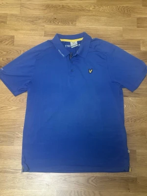 Blå kortärmad t-shirt Lyle & Scott - Blå t-shirt från Lyle & Scott med klassisk pikékrage och knappslå. Ikonisk gul logga på bröstet och diskreta logotyper på krage och ärm. Tillverkad i mjuk bomull för en skön känsla och normal passform.