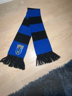 Blåsvart halsduk Sirius IF - Snygg halsduk i blått och svart med breda ränder och fransar i ändarna. Halsduken har Sirius IF:s klubbmärke broderat på ena sidan. Perfekt för att visa ditt stöd på matchen eller som accessoar till vardagslooken.