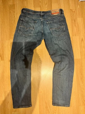 Blå diesel jeans - Säljer ett par klassiska blå jeans från diesel med raka ben och två bakfickor med ikonisk V-söm. Jeansen har en vintage look och är tillverkade i slitstark denim. Perfekta för dig som gillar tidlös stil och vill ha ett par jeans som funkar till allt.