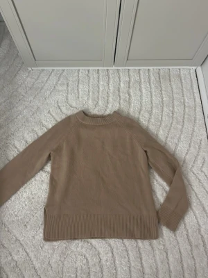 Beige stickad tröja med slits från hm  - En stilren beige stickad tröja med rund halsringning och långa ärmar. Tröjan har ribbade muddar vid ärmslut och nederkant samt en liten slits i sidan för extra detalj. Perfekt för lager på lager och enkel att matcha med olika outfits.