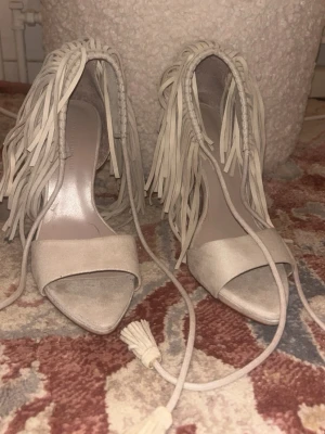 Beige pumps med fransar från Zara - Säljer ett par beige pumps från Zara med öppen tå, höga klackar och dekorativa fransar längs kanten. Skorna har snörning med tofsar och är tillverkade i mockaimitation. Perfekta för dig som vill sticka ut med en unik detalj!