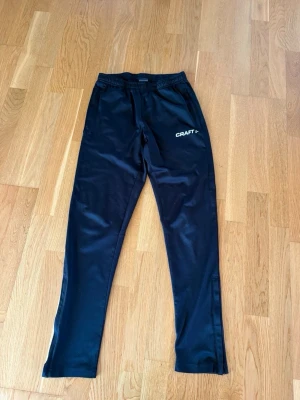 Svarta träningsbyxor från Craft - Säljer ett par svarta träningsbyxor från Craft med resår i midjan och logga på vänster lår. Byxorna har raka ben och är tillverkade i ett lätt, funktionsmaterial som passar perfekt till löpning eller gym. Nästan aldrig använda 