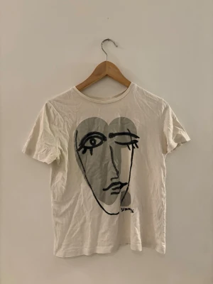 Vit t-shirt med konstnärligt ansikte - Vit t-shirt i bomull med ett stort grafiskt tryck av ett abstrakt ansikte i svart på framsidan. T-shirten har rund hals och korta ärmar. Perfekt för dig som gillar unik och konstnärlig stil.