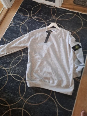 Grå sweatshirt från Stone Island - Säljer en stilren grå sweatshirt från Stone Island med klassisk rund hals och ribbade muddar. Tröjan har den ikoniska loggan på vänster ärm och är tillverkad i mjukt bomullsmaterial. Perfekt för en avslappnad och trendig look.
