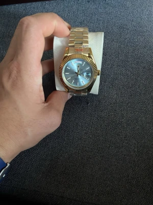 Guldfärgad klocka med blå urtavla - Stilren klocka i guldfärgat metallarmband och boett. Urtavlan är blå med tydliga indexmarkeringar och datumvisning. Klassisk design med räfflad bezel och länkarmband som ger en exklusiv känsla.