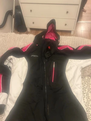 Svart och vit skidjacka från Phenix - Snygg svart och vit skidjacka från Phenix med rosa detaljer och dragkedja. Jackan har huva, bröstficka med dragkedja och sportig design. Perfekt för skidåkning och vinteraktiviteter. Fodrad insida i rosa för extra stilpoäng.